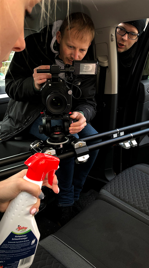 Behind-the-Scenes von Autopolsterreinigungs-Werbespot mit Frau die Reinigungsflasche hält während zwei Deichblick-Crewmitglieder filmen.