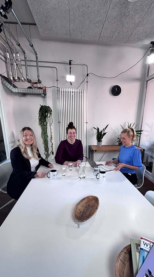 Dynamisch aus leichter Schrägperspektive fotografiert: Tabea Herrera, Kristina Adams und Alexandra von Lingen am weißen Interviewtisch mit Wasserkaraffe, Kaffeetassen und Laptop, mit einladendem Lächeln in die Kamera blickend.