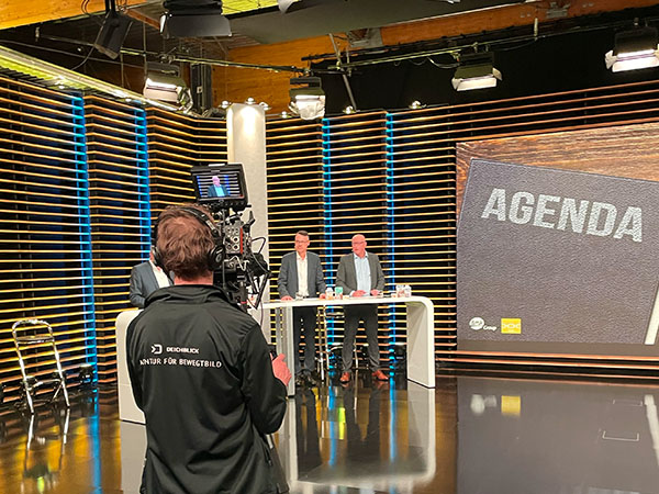 Blick durch ein Regiefenster auf eine Frau, teilweise verdeckt durch Spiegelungen von Studiobeleuchtung, Flaschen und einem Monitordisplay – authentischer Einblick in den Regieraum einer Produktion.