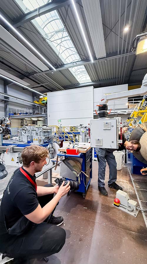 Joshua Hartman von Deichblick kniet in HighVolt Halle und fotografiert Mitarbeiter bei der Arbeit. Mitarbeiter steht hinter Box an Metallarm befestigt, schaut seitlich hervor.